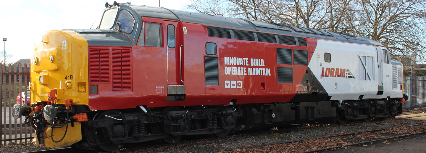 A Loram train sits outside on tracks in front of several trees.
