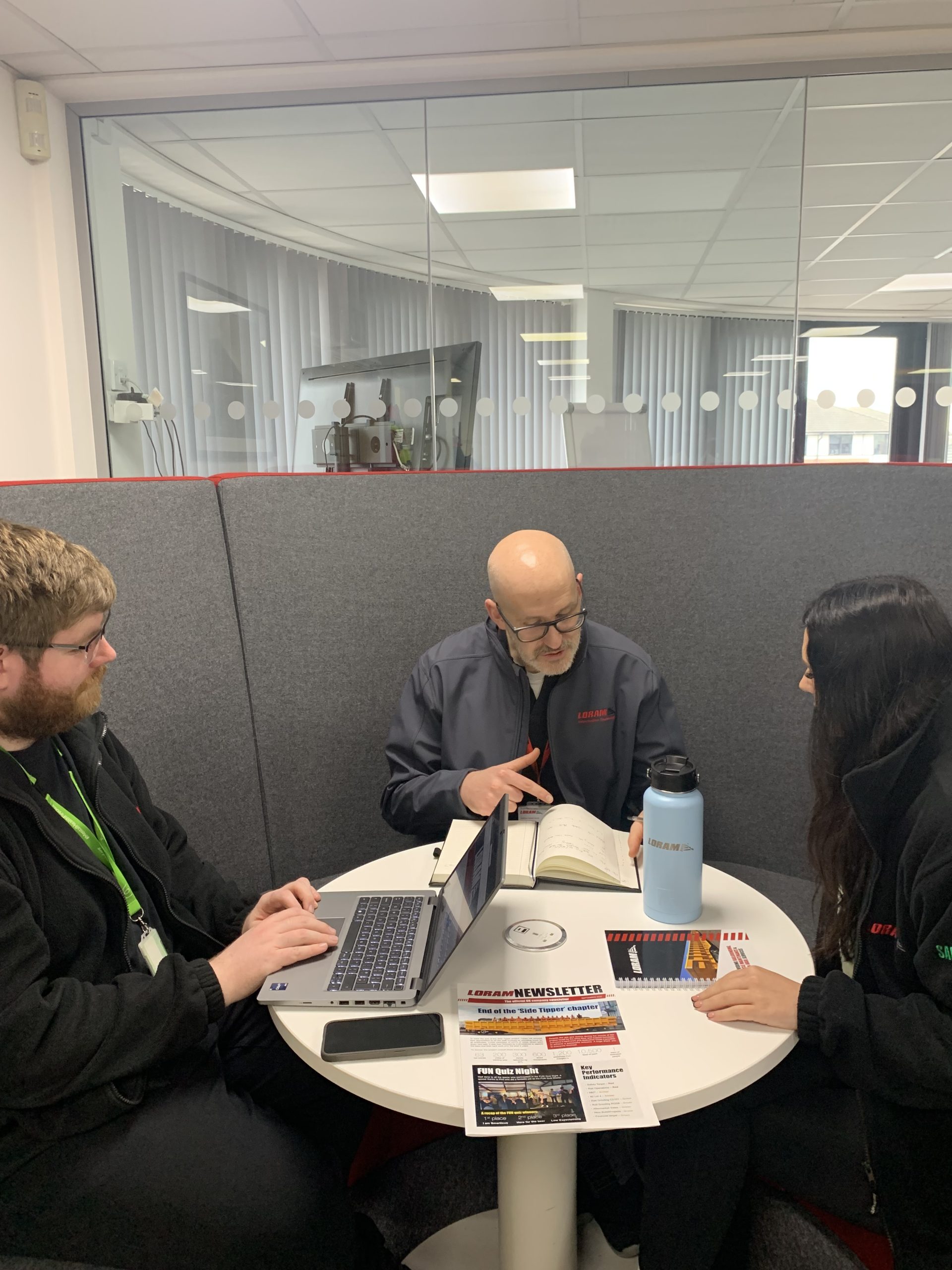 Three Loram colleagues in a modern office collaborate around a small table with a laptop, notebook, and newsletter.