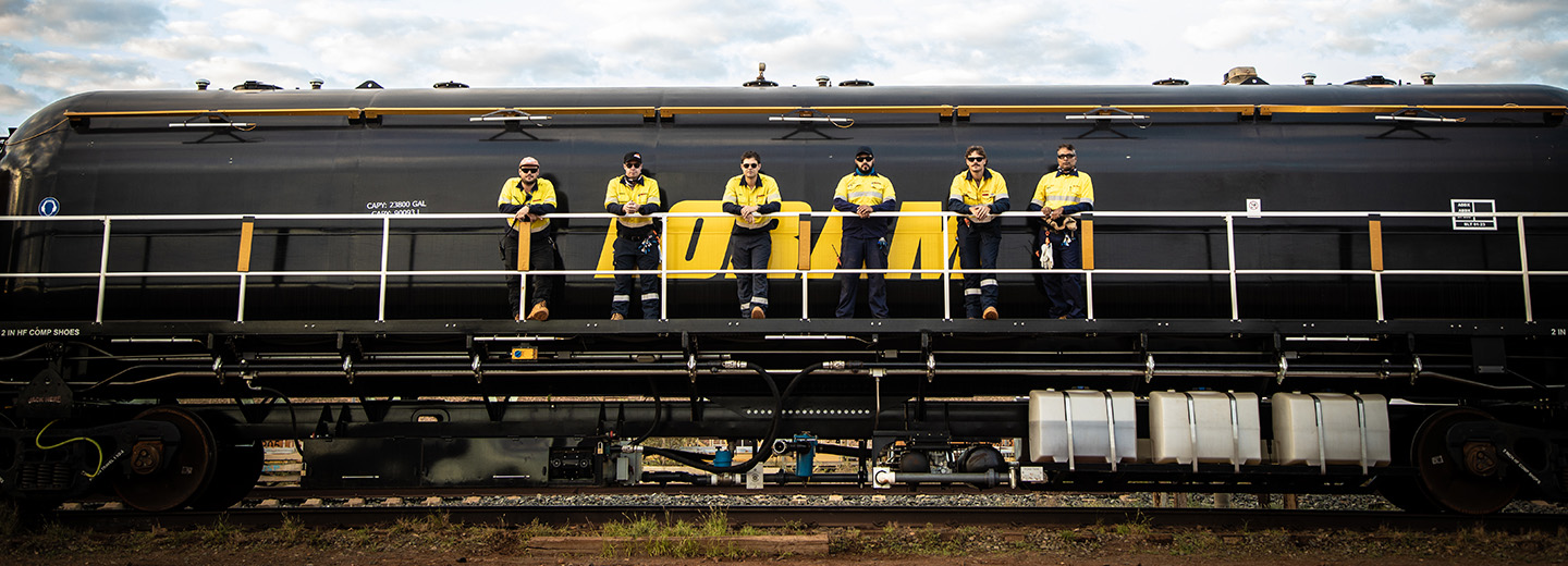 Six people in yellow shirts lean against a railing in front of a train.